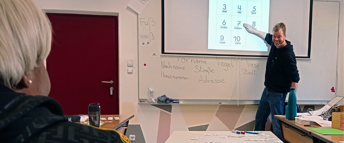 Lehrer Mathias zeigt auf der Tafel auf ein Nummernfeld, um Zahlen auf Deutsch zu unterrichten